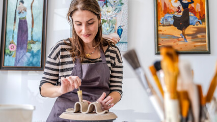 A close-up shot of a middle-aged woman artist shaping clay with sculpting tools at a table in an...