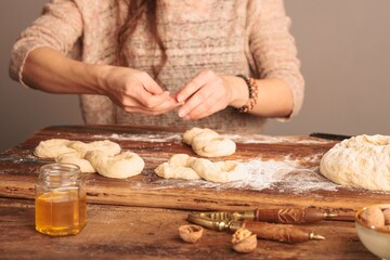 A woman forms the traditional figure-eight shape of Mucenici from dough on a rustic wooden table. Walnuts, honey, and a nutcracker complete the sweet treat preparation