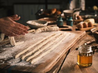 A person rolls out dough on a floured wooden board, preparing Mucenici in a warm, rustic kitchen setting. Ingredients like honey and eggs are nearby, ready for baking the traditional treats