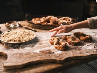 Delicately shaped Mucenici are arranged on a floured wooden board, freshly baked. A hand reaches toward the pastries, with a plate of chopped walnuts and a tray of more desserts behind