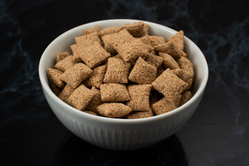 A white bowl filled with crisp, whole grain cereal sits on a dark surface, highlighting the texture and shape of the pieces. This setup is perfect for breakfast or a midday snack.