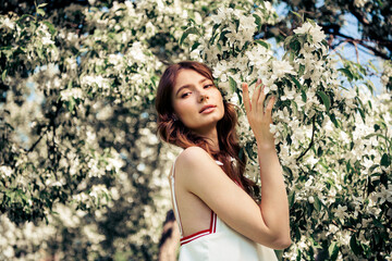 Pretty woman wearing a white summer dress outdoors among blooming flowers during daylight,...