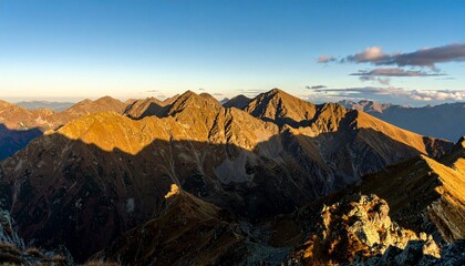 Naklejka premium Panoramic view of rugged mountain peaks in autumn, golden sunlight casting warm glow over rocky ridges, vast landscape with dramatic shadows