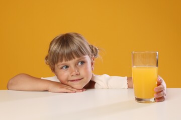 Cute little girl reaching for glass of juice at white table against orange background