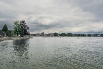Serene lakeside view with calm water reflecting overcast sky and lush greenery, creating a tranquil...