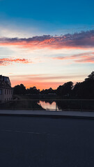 Obraz premium Serene sunset over calm river with vibrant colors reflecting on water, framed by trees and buildings, creating a tranquil evening atmosphere