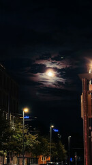 Nighttime urban scene featuring a bright full moon illuminating clouds above a quiet street with...