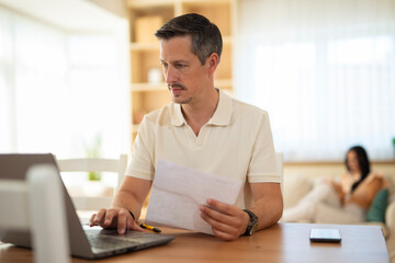 Fototapeta premium Focused man working on a laptop while holding documents at home, with a woman relaxing in the background. 