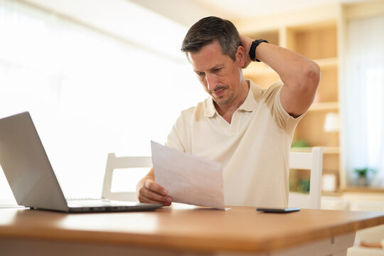 Worried man looking at document while working on laptop at home, feeling concerned about financial situation.
