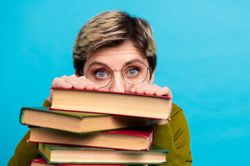Young woman with short hair wearing glasses behind a pile of book on a blue background, expressing...
