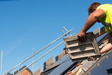 Roofer carefully places roof tiles on a sloped surface while standing on top of a building under a...