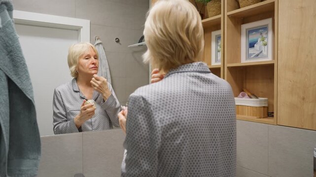 A senior lady in pajamas applying nourishing face cream while looking in a mirror. Her beauty regimen includes moisturizing and anti-wrinkle treatment for skin rejuvenation and hydration.
