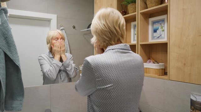 Mature woman spreads nourishing cream over her face and neck, transitioning into a self-massage. This beauty treatment uses lifting techniques for better skin absorption and a firming effect.