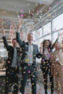 Blurred background of people in business suits celebrating in an office, throwing confetti and holding party streamers. Concept of corporate celebration, team success, and joyful workplace moments.