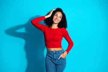 Confident young woman in a red top and jeans posing against a bright blue background, embracing a...
