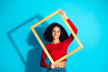 Beautiful young woman holding a wooden frame against a bright blue background, showcasing stylish...