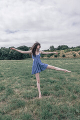 Young woman with long curly brown hair wearing a sleeveless blue dress, standing barefoot on grass in an open field, balancing gracefully with one leg extended and arms outstretched