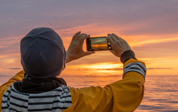 Asnelles-Sur-Mer, France - 08 08 2025: View of the jetty with a mature woman in a yellow raincoat and a hat taking pictures with a smartphone of the sea and the sky at sunset - Powered by Adobe