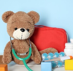 Pediatrician concept. Toy bear, cubes, first aid kit, pills, thermometer and stethoscope on white wooden table against light blue background, closeup