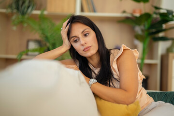Sad woman sitting on couch at home, leaning on hand and looking away with pensive expression,...