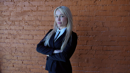 Confident young businesswoman in a suit poses with crossed arms in front of a brick wall, exuding...
