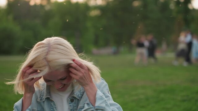 beautiful smiley woman with white bob hair seated on grass laughing hard while watching blurred people playing in park, denim jacket, summer evening light casting warm glow, carefree social moment