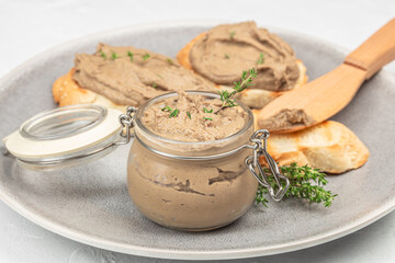 Glass jar of creamy pate garnished with fresh thyme, served with toasted bread slices on a gray plate wooden spreader, close-up view