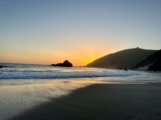 Golden sunset over the Pacific Ocean with waves washing onto the sandy beach and rugged cliffs in the background.