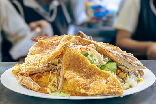 Ecuadorian hornado, a traditional Ecuadorian dish, oven-cooked until achieving juicy meat and crispy skin, served with toasted corn, llapingachos, fresh salad, avocado, and white hominy.