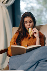 Surprised woman reading a book while eating a sandwich in cozy indoor setting, with natural light...