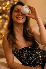 A beautiful young woman stands next to a Christmas tree. A gorgeous lady in a black sparkling dress with long dark hair is decorating the tree. A New Year's background with a Christmas tree and happy 