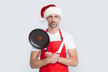 mature christmas man in red santa hat and apron hold frying pan and kitchen knife