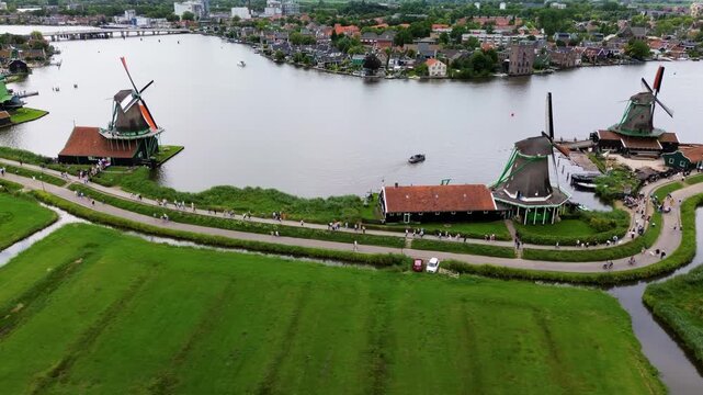 Drone view of traditional Dutch windmills along the river, surrounded by green fields, canals, and tourists walking on scenic paths in Zaanse Schans. Picturesque Dutch windmills by the river.