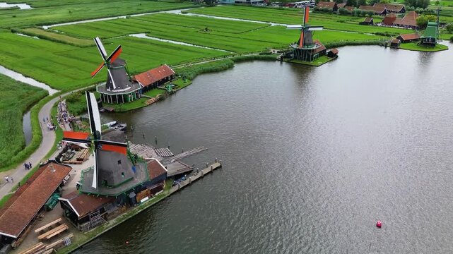 Drone view of traditional Dutch windmills along the river, surrounded by green fields, canals, and tourists walking on scenic paths in Zaanse Schans. Picturesque Dutch windmills by the river.