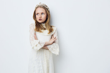Portrait of a young girl in a vintage white dress with a tiara, looking thoughtful against a light...