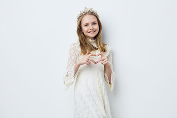 Young girl in a white dress and crown making heart shape with hands, joyful expression, minimalist...