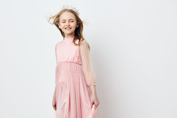 A joyful young girl in a light pink dress smiles while standing against a clean white background,...