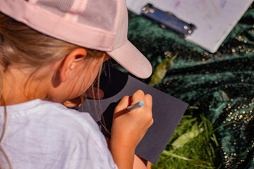 Young girl wearing a pink cap is focused on drawing with a pen on a black sheet of paper,...