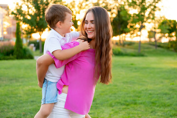 Fototapeta premium mom and baby playing on a green lawn in the summer, having fun together, mother and son walking together on a summer evening, with the baby on her neck