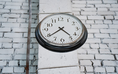 Wall mounted round analog clock showing time on white brick industrial building background with metal pipe detail representing punctuality accuracy and time management concept
