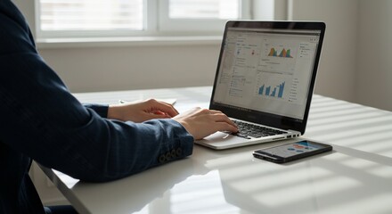 Fototapeta premium Business analytics dashboard on laptop with KPI metrics and charts; hands typing at modern office desk with smartphone and window light for data-driven marketing and finance