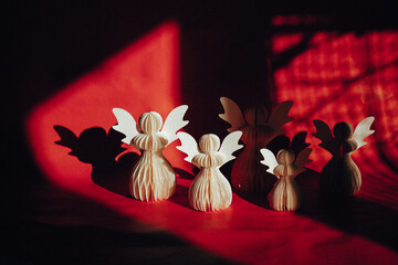 Paper angel figurines are arranged on red background in the lighting, creating a striking contrast composition. A group of paper figurines are illuminated by a dramatic light, casting long shadows.