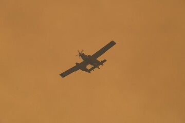 Firefighting aircraft dropping water over burning forest landscape, casting dramatic silhouette against smoke filled sky in leon, spain