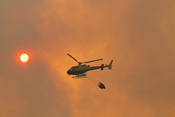 Helicopter battling wildfire, dropping water against a backdrop of a smoky orange sunset, highlighting the urgent fight against nature's fury