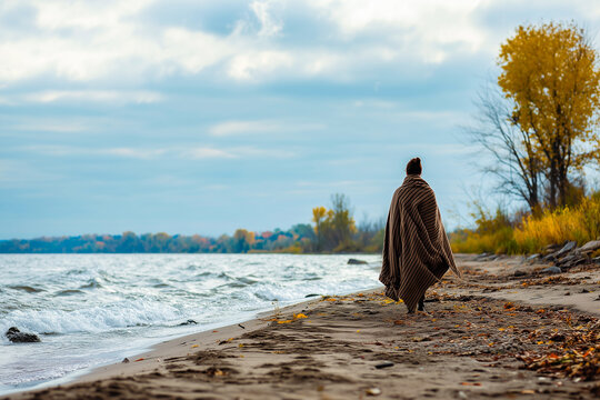 Tranquil scene of a woman wrapped in a blanket walking along the shore on a breezy autumn day - Powered by Adobe