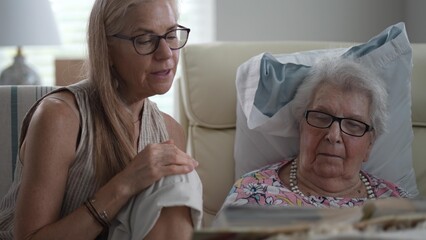 In a senior living facility, a daughter and her elderly mother laugh together while reminiscing over a photo album filled with cherished memories.