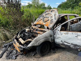 Heavily burnt car with its hood open, exposing a charred and rusted engine, abandoned on a rural...