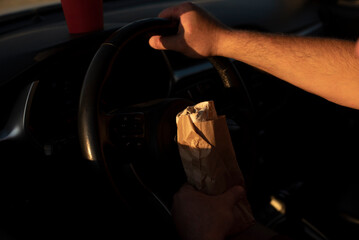 A hand grips a steering wheel while holding a brown paper bag. The scene is illuminated by warm...