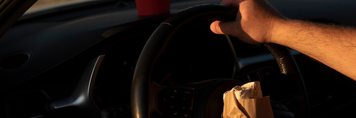 A hand gripping a steering wheel inside a car. A brown paper bag rests on the dashboard. The scene...