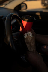 A person holds a red coffee cup and a wrapped sandwich while driving. The scene emphasizes...